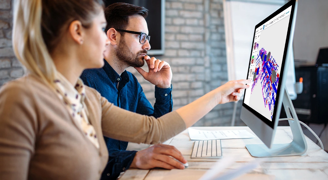 Two professionals collaborate in an office, reviewing a detailed 3D building model on a monitor using Trimble software.