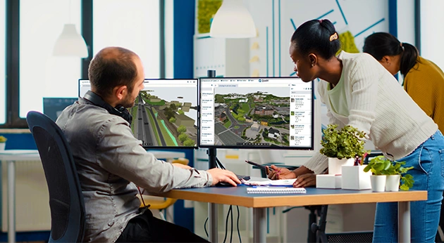 Two professionals in an office collaborate using Trimble Quadri software on dual monitors to analyze a 3D road and terrain model.