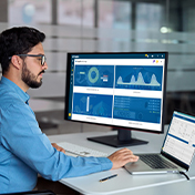 A man with a beard and glasses sits at a desk, looking at a computer monitor displaying graphs and data from Supplier Xchange.