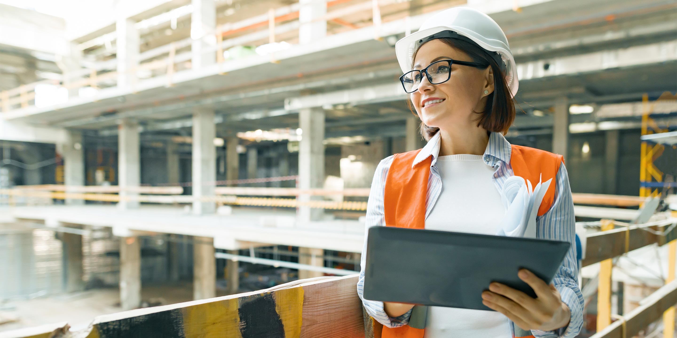 A woman in a hard hat and orange vest smiles while holding a tablet and papers at a construction site.