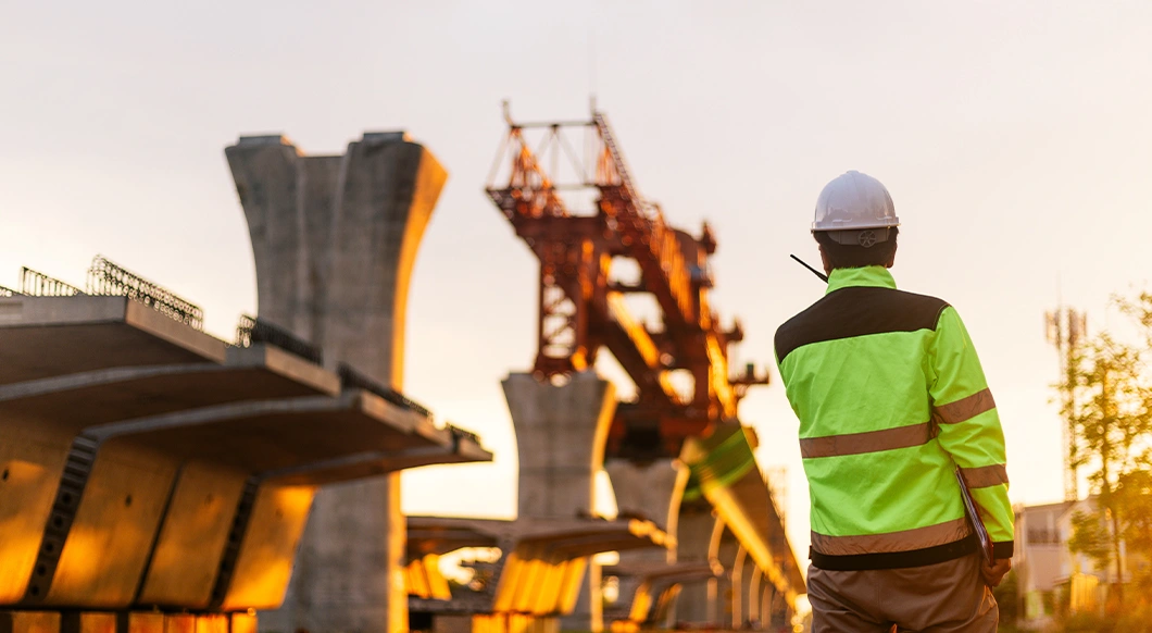 A civil engineer in a hi-vis vest overlooks a bridge construction site with a red launching gantry and concrete segments at sunset.