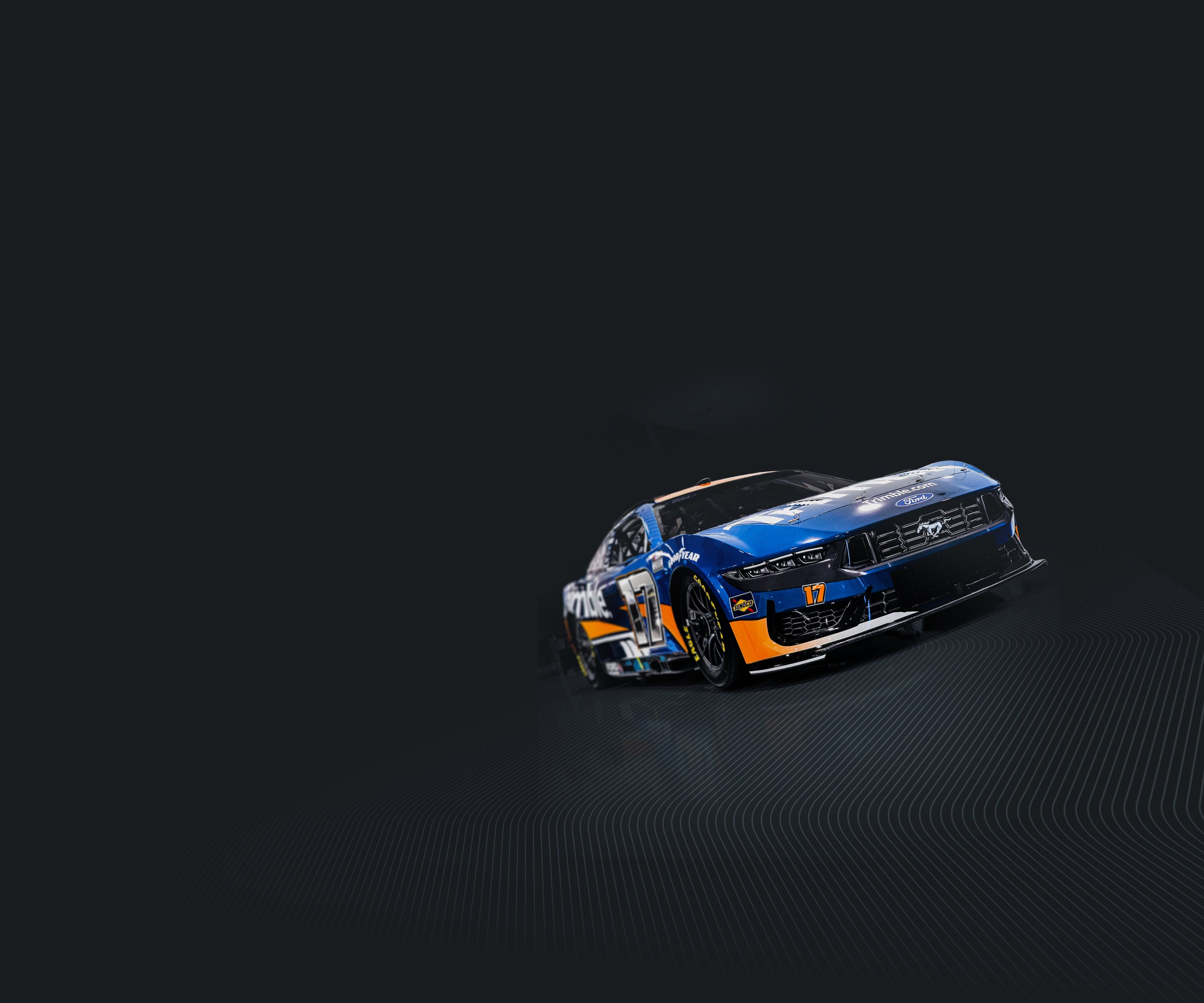 A blue and orange No. 17 Trimble Ford Mustang race car driven by Chris Buescher for RFK Racing, set against a dark, minimalist background.