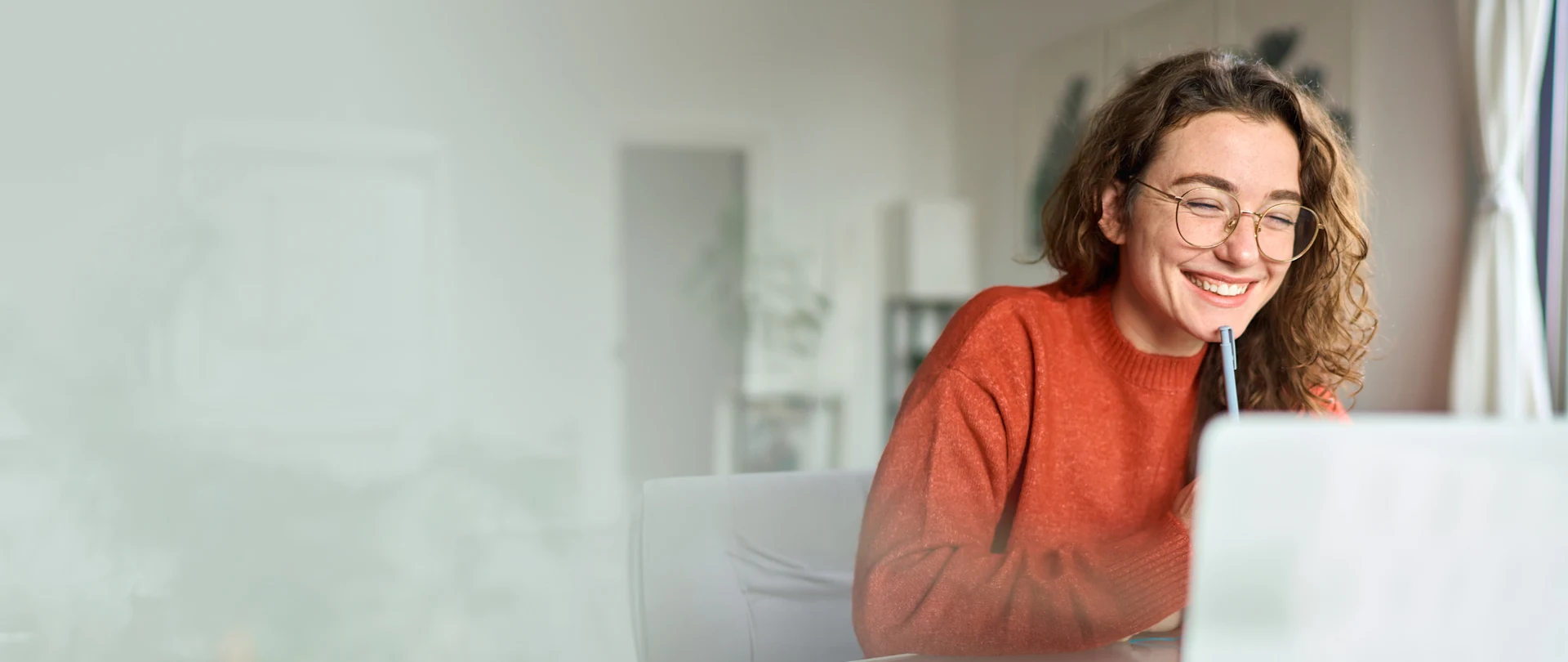 A smiling woman with glasses and curly hair in an orange sweater sits at a desk with a laptop and pen in a bright, modern room.