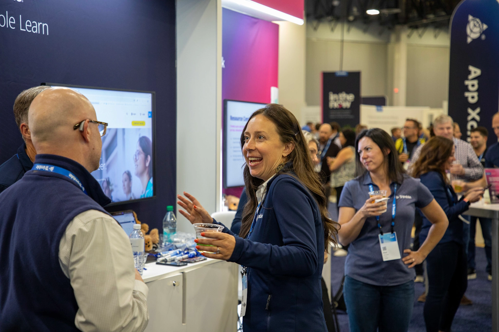 A smiling woman holding a drink chats with a man in a vest at a busy trade show booth. Other attendees network in the background near a display screen showing digital content.