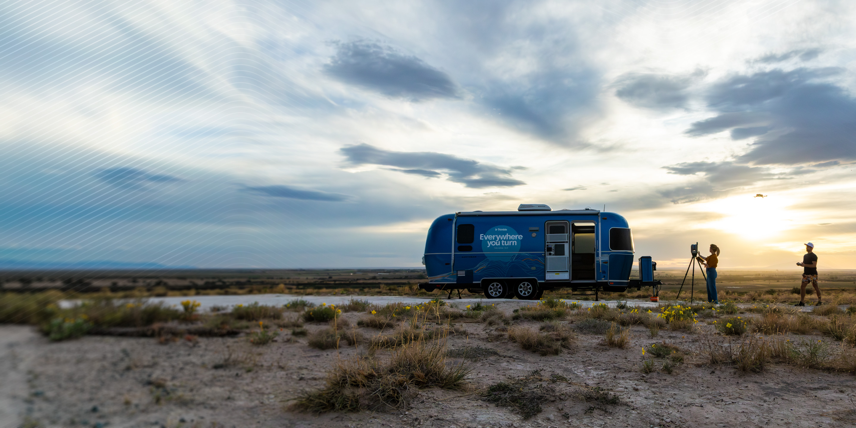 A blue Trimble Airstream trailer at sunset. Two professionals use a surveying scanner on a tripod and a drone in an open landscape.