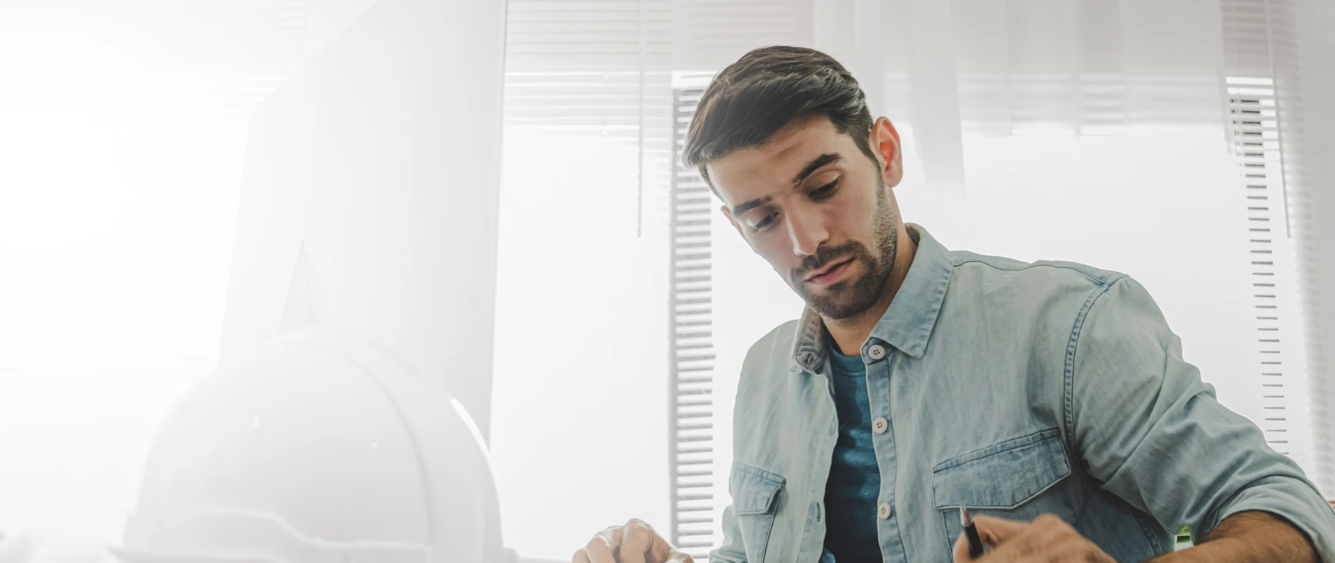 A man in a denim shirt examines blueprints on a drafting table next to a white hard hat in a sunlit office.