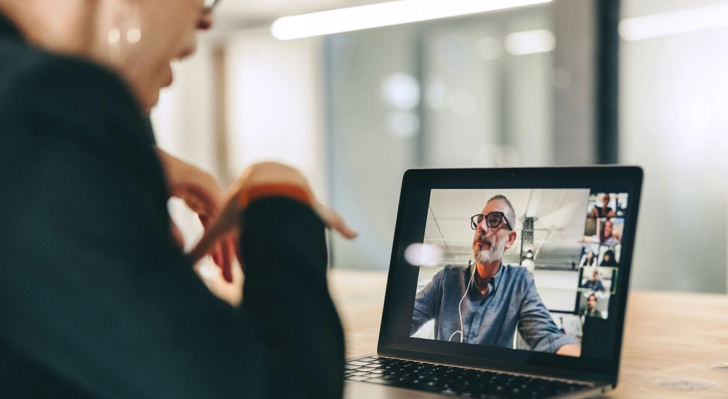 Woman in a professional office participating in a video call on a laptop with a bearded man and other colleagues.