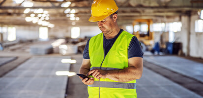 A construction worker in a yellow hard hat and safety vest uses a smartphone in a warehouse, representing Trimble's digital field solutions.