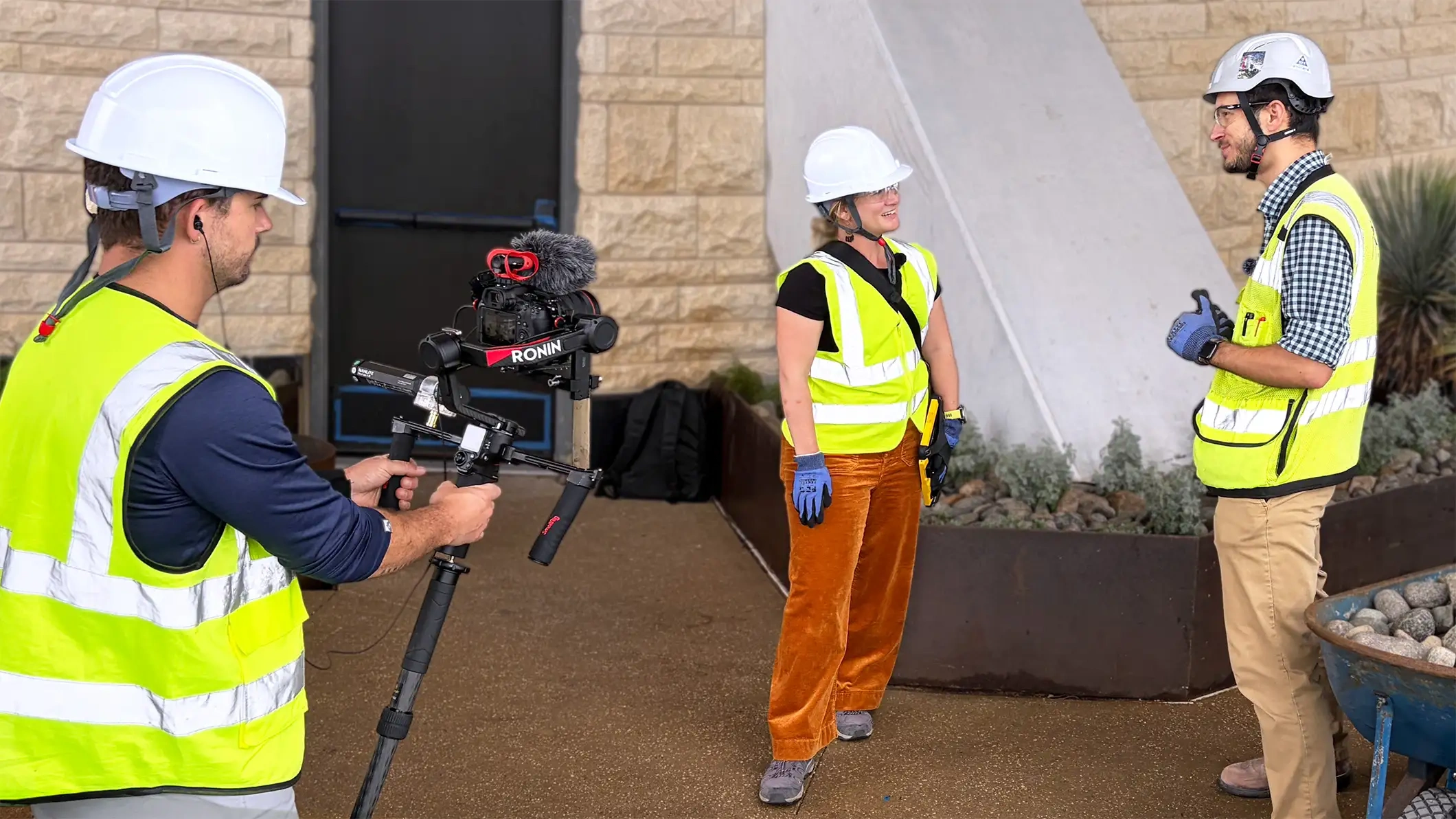 Auf einer Baustelle filmt ein Videograf mit einem Gimbal zwei Fachleute, die Schutzhelme und Sicherheitswesten mit Trimble-Logo tragen.