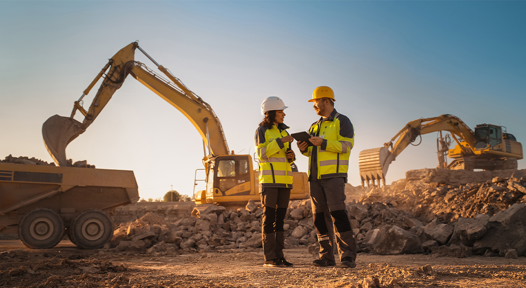 Two construction workers review a tablet at a job site with excavators and a dump truck during a golden sunset.