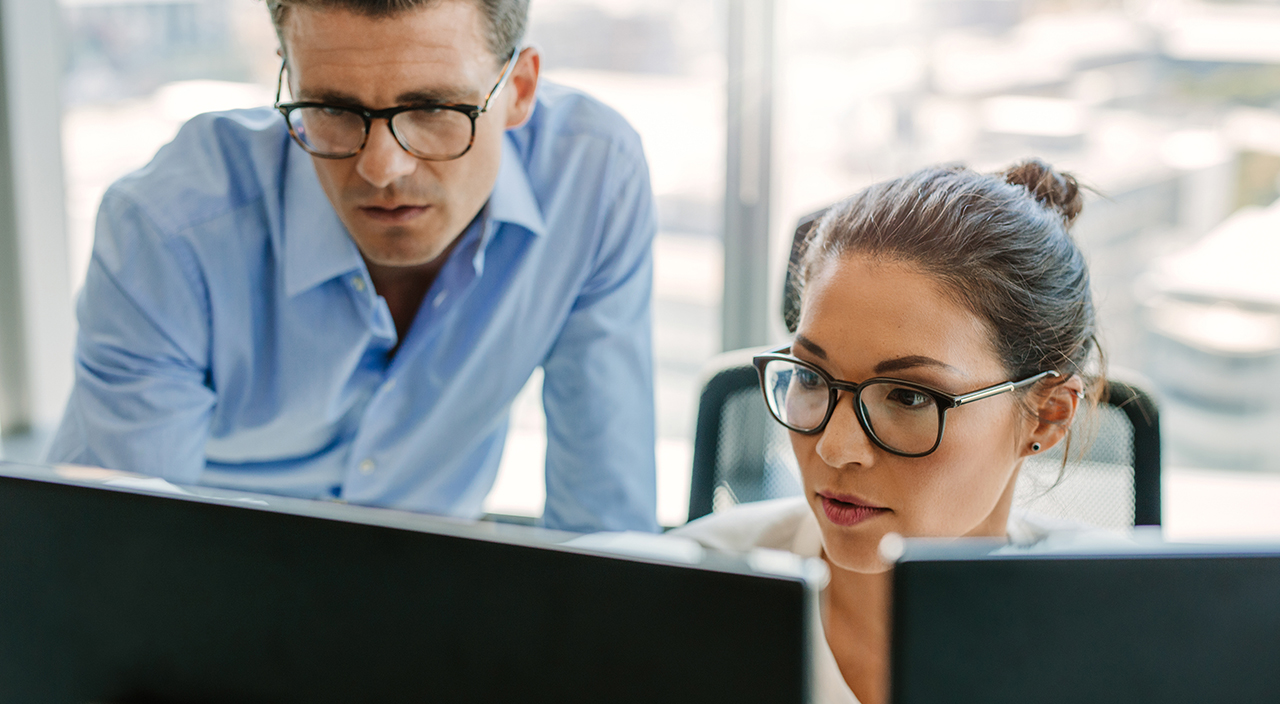 A man and a woman with glasses collaborate while looking at computer monitors in a bright, modern office setting.