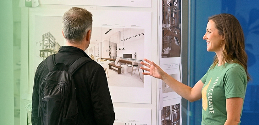 In a modern showroom, a woman wearing an "IGNITE CREATIVE" t-shirt gestures toward Trimble's SketchUp architectural designs while presenting to a man.