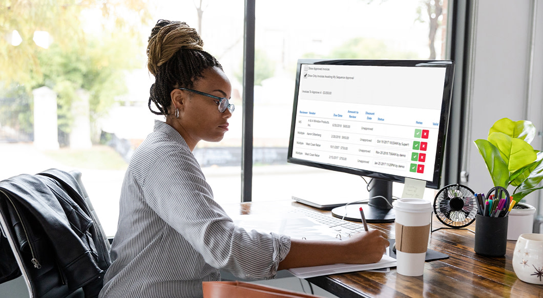 A woman at a desk reviews Trimble Viewpoint's "Invoices To Approve" dashboard on a computer monitor while writing on a document.