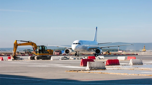 Construction barriers and heavy machinery at airport runway with airplane in background