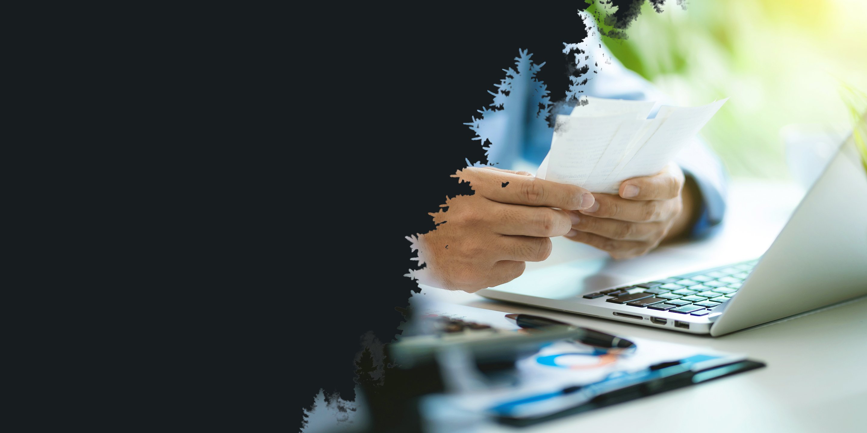 A person holds and examines papers near an open laptop on a desk. The scene conveys focus and concentration, with a blurred green background.
