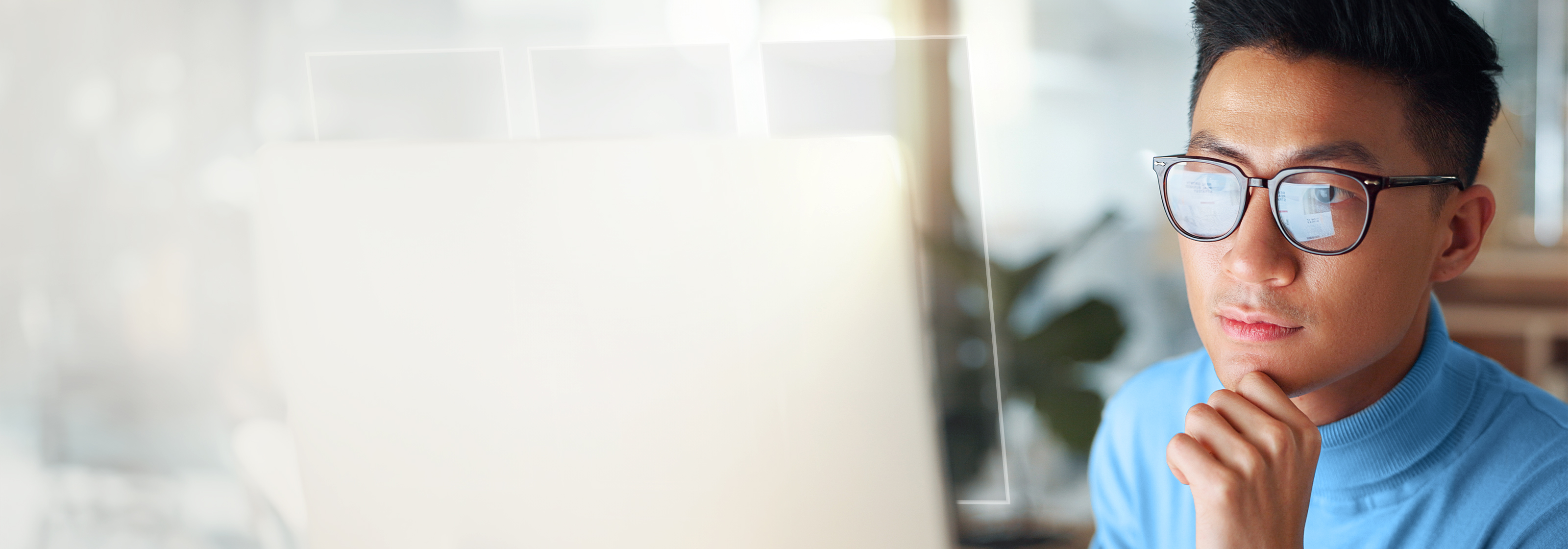 A close-up of a man wearing dark-rimmed glasses and a blue turtleneck, looking intently at a brightly lit, blurred laptop screen, with his hand resting under his chin.