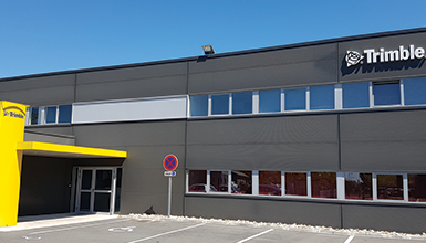 A low-angle exterior shot of a modern, dark-gray building with a yellow sign for Trimble in front.
