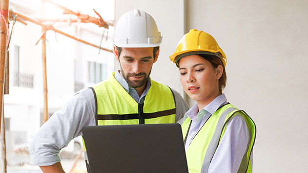 A male and female construction worker in hard hats and safety vests collaborating on a laptop at a job site.