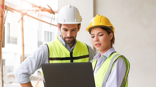 A male and female construction worker in hard hats and safety vests collaborating on a laptop at a job site.