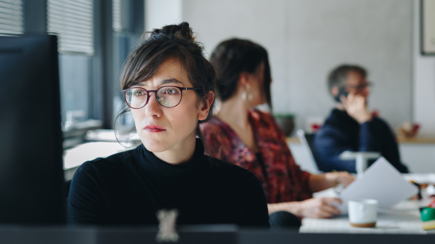 A woman with glasses and her hair in a bun intently looks at a computer screen in a bright office environment.