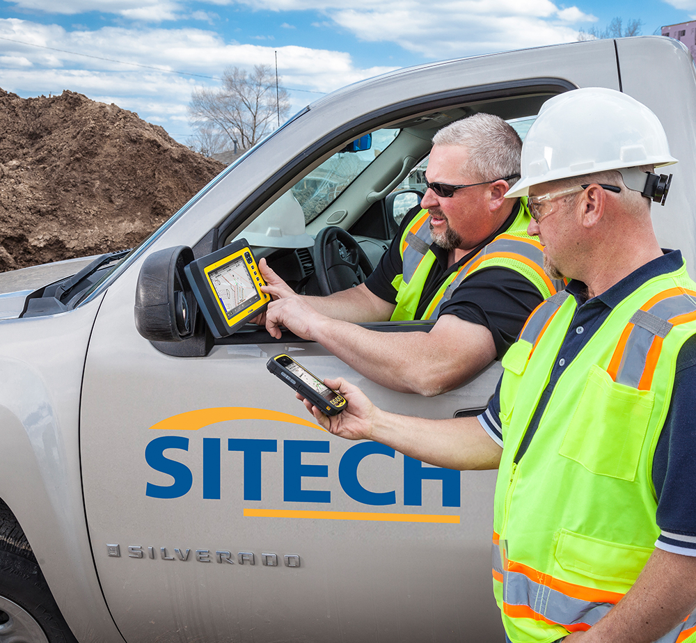 Two construction workers in vests and hard hats look at a device mounted to a SITECH truck window.