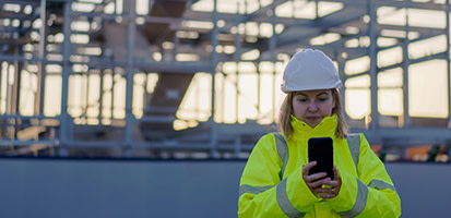 A woman in a white hard hat and yellow high-visibility jacket uses a smartphone at a construction site with a steel frame structure.