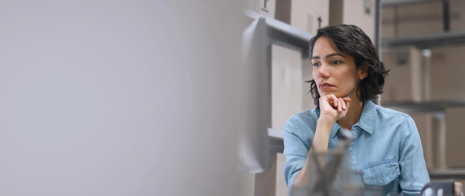 A woman thoughtfully looks at a computer screen in a warehouse office, with shelves of boxed inventory in the background.