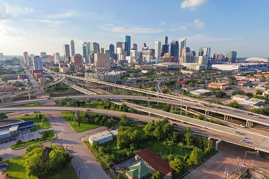 Aerial view of the downtown Houston skyline with complex highway interchanges in the foreground under a clear sky.