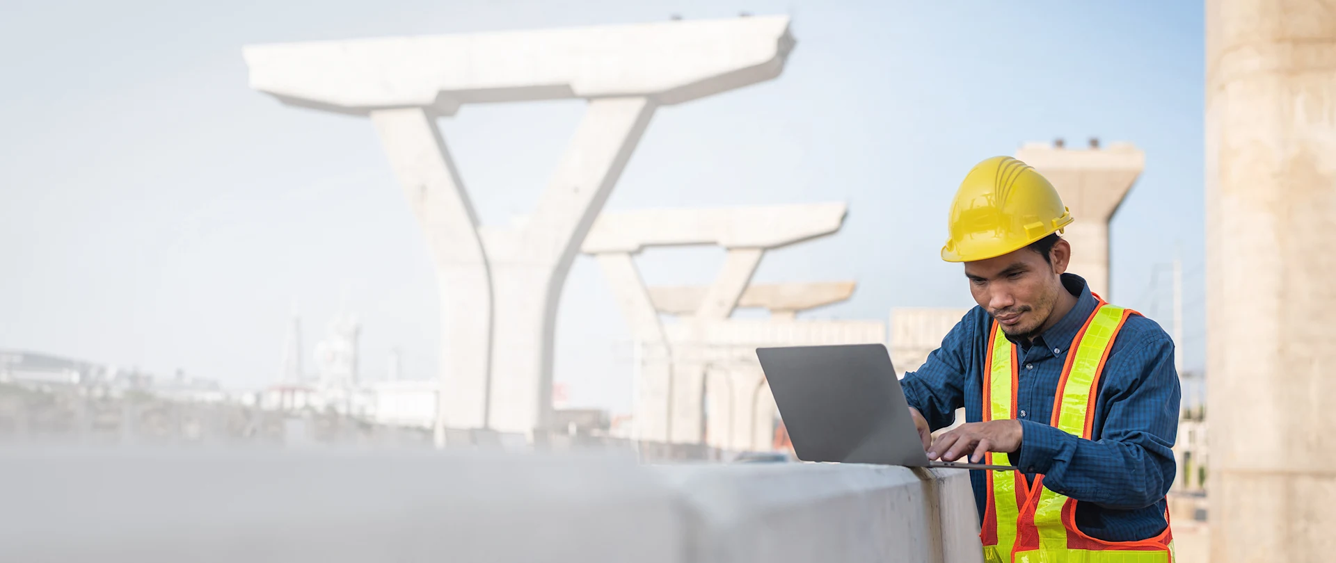 A construction engineer in safety gear uses a laptop at a bridge construction site in Nong Khai, Thailand.