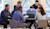 A diverse group of seven people is gathered around a table, looking at a laptop and a tablet.