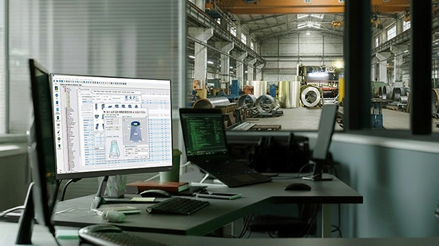 A desk with two computer screens displays Trimble FabShop software with a view of a HVAC fabrication workshop behind it.