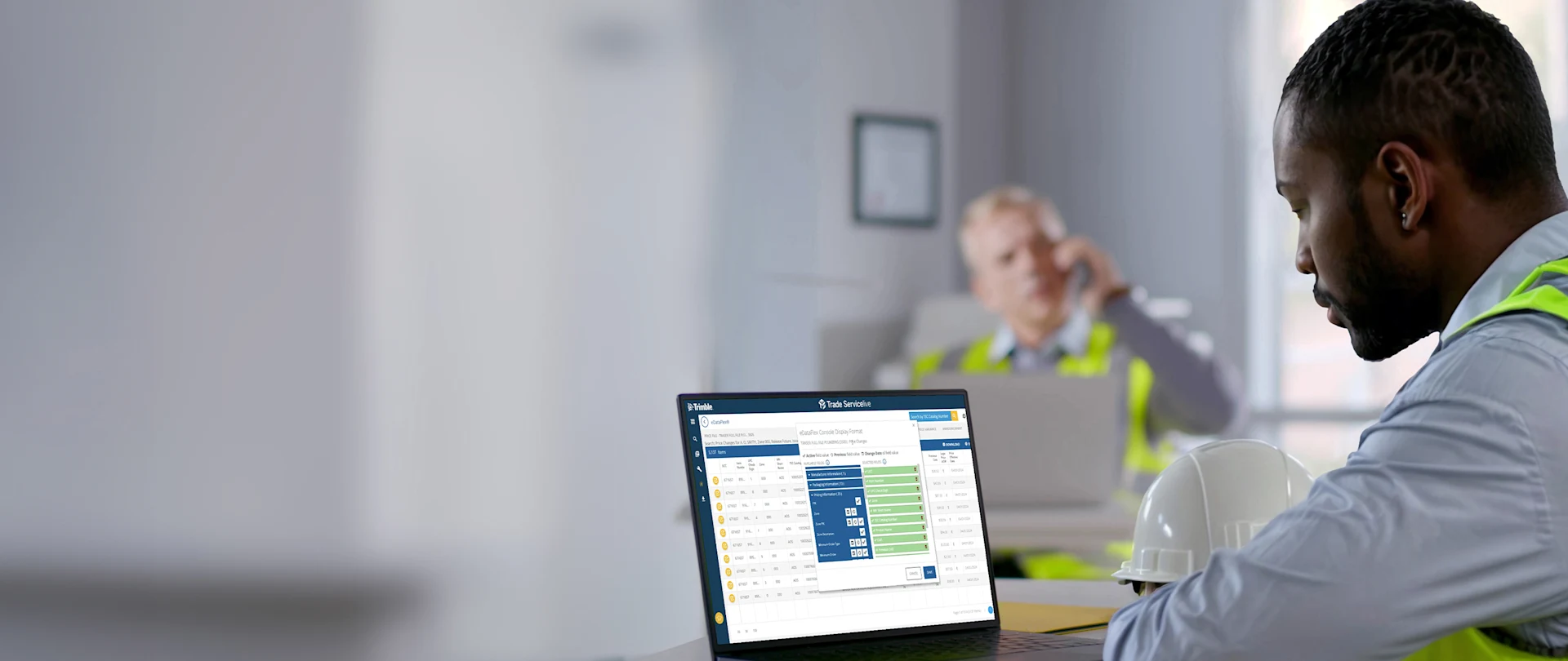 A Black man in a safety vest uses Trimble Trade ServiceLive eDataFlex on a laptop next to a white hard hat in an office.