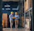 Two men in yellow hard hats discuss inventory in a warehouse aisle; one holds a tablet and the other a yellow folder.