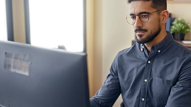 A man with glasses and a beard wearing a blue button-down shirt looks intently at a computer monitor in a bright office setting.