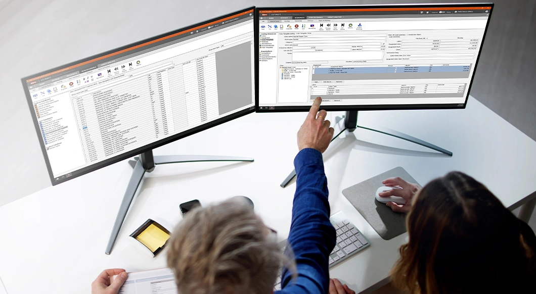 A man and woman use HCSS Estimate construction software on dual monitors in a modern office to manage project data and analyze costs.