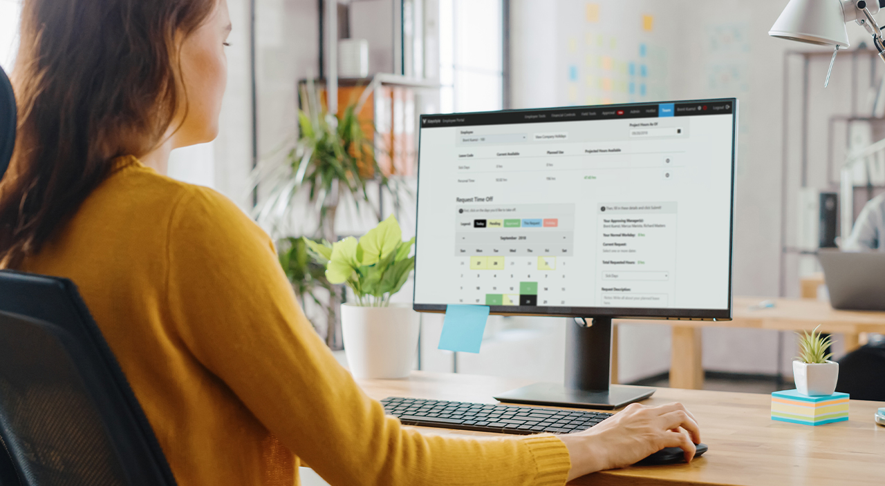 A woman works at a desk using the Trimble Viewpoint employee portal on her monitor to submit a time-off request on a digital calendar.
