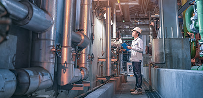 An industrial engineer in a hard hat and safety gear inspects large pipes and valves in a facility, recording data on a blue clipboard.