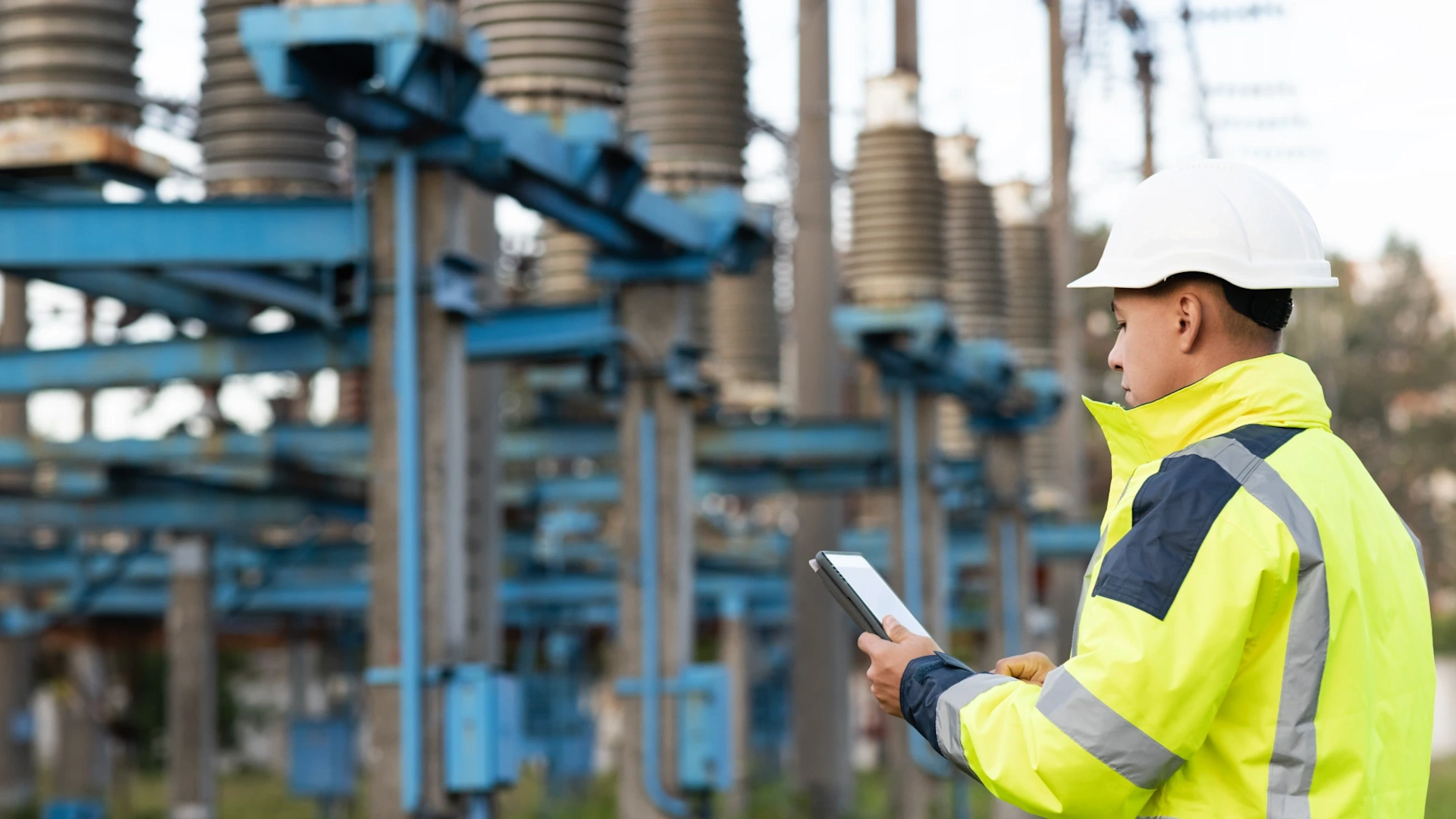 An AECO professional in a hard hat and hi-vis jacket uses a tablet for digital inspection at an electrical substation; ribbed insulators and blue structures are visible.