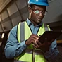 An African American field worker in a blue hard hat and high-visibility vest uses a tablet at an industrial job site.