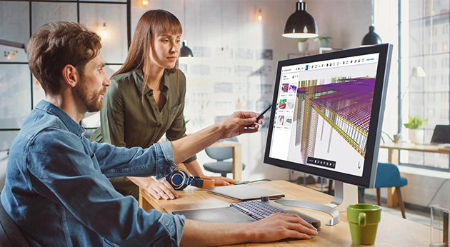 Two colleagues collaborate in a modern office, reviewing a detailed 3D structural building model on a monitor using Trimble Tekla Structures software.
