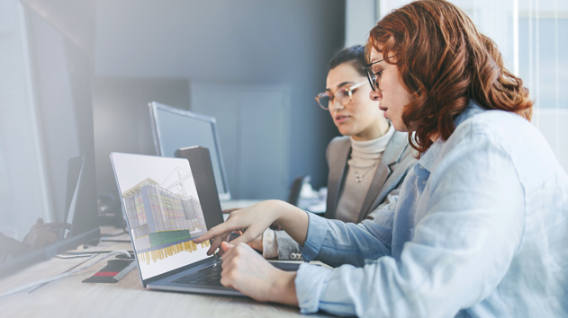 Two women collaborate in an office, reviewing a 3D building model on a laptop using Trimble Tekla Structures software.