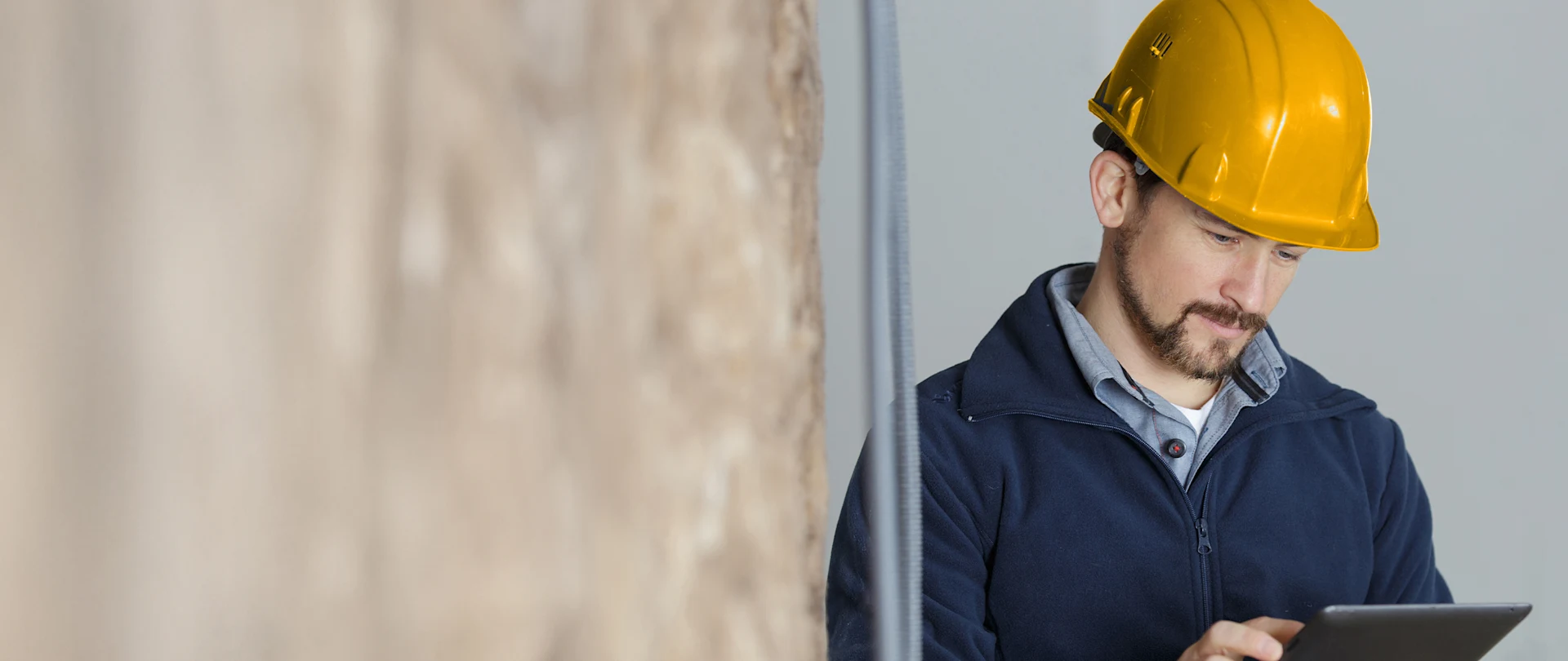 A construction professional in a yellow hard hat and navy fleece using a tablet indoors near a textured wall.