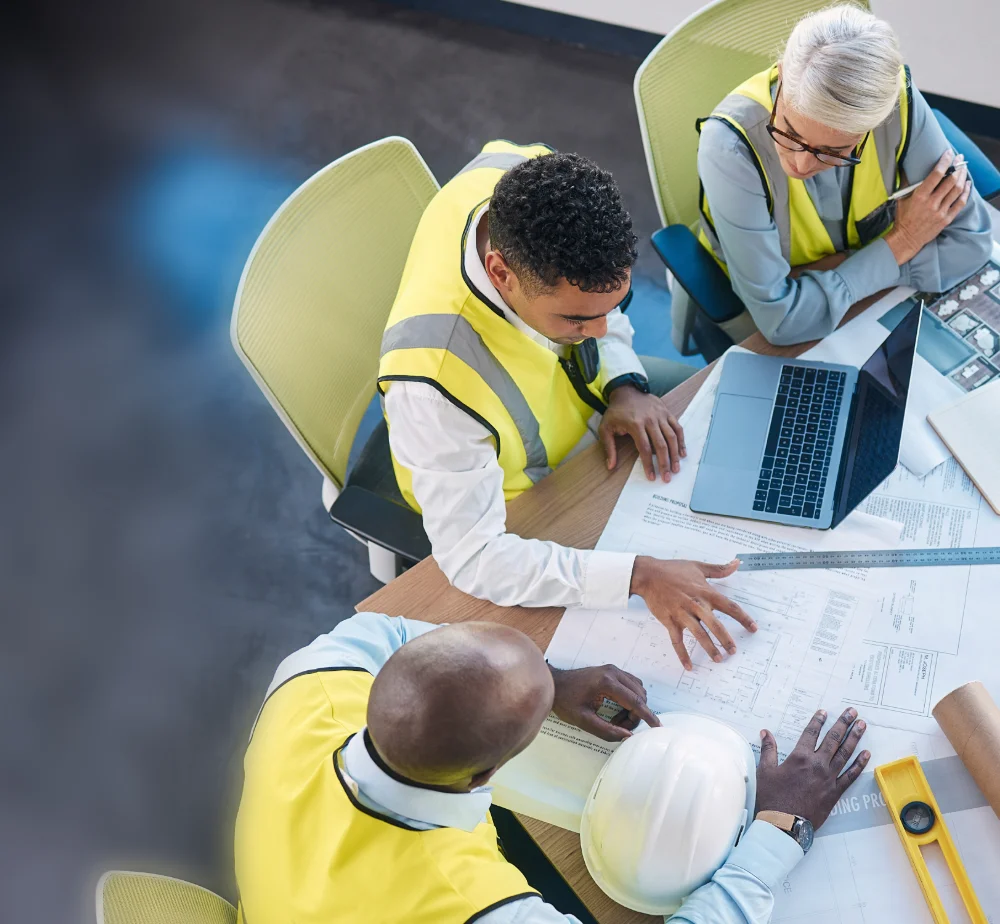 Three AEC professionals in yellow vests review 'BUILDING PROPOSAL' blueprints. An Apple MacBook Pro, white hard hat, and yellow spirit level are on the table.