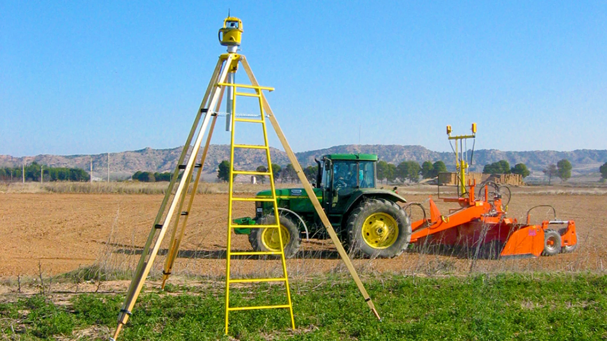 A Trimble Spectra Precision GL700 laser transmitter on a tripod guides a tractor leveling a field with an orange scraper in the background.