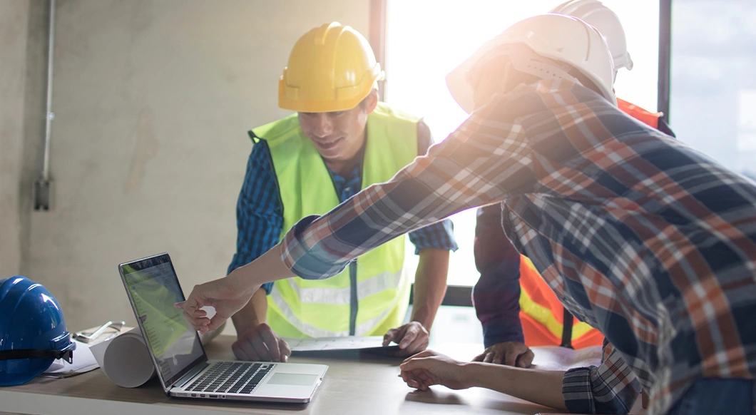 Construction workers in safety gear review project data on a laptop running Trimble software at an indoor building site.