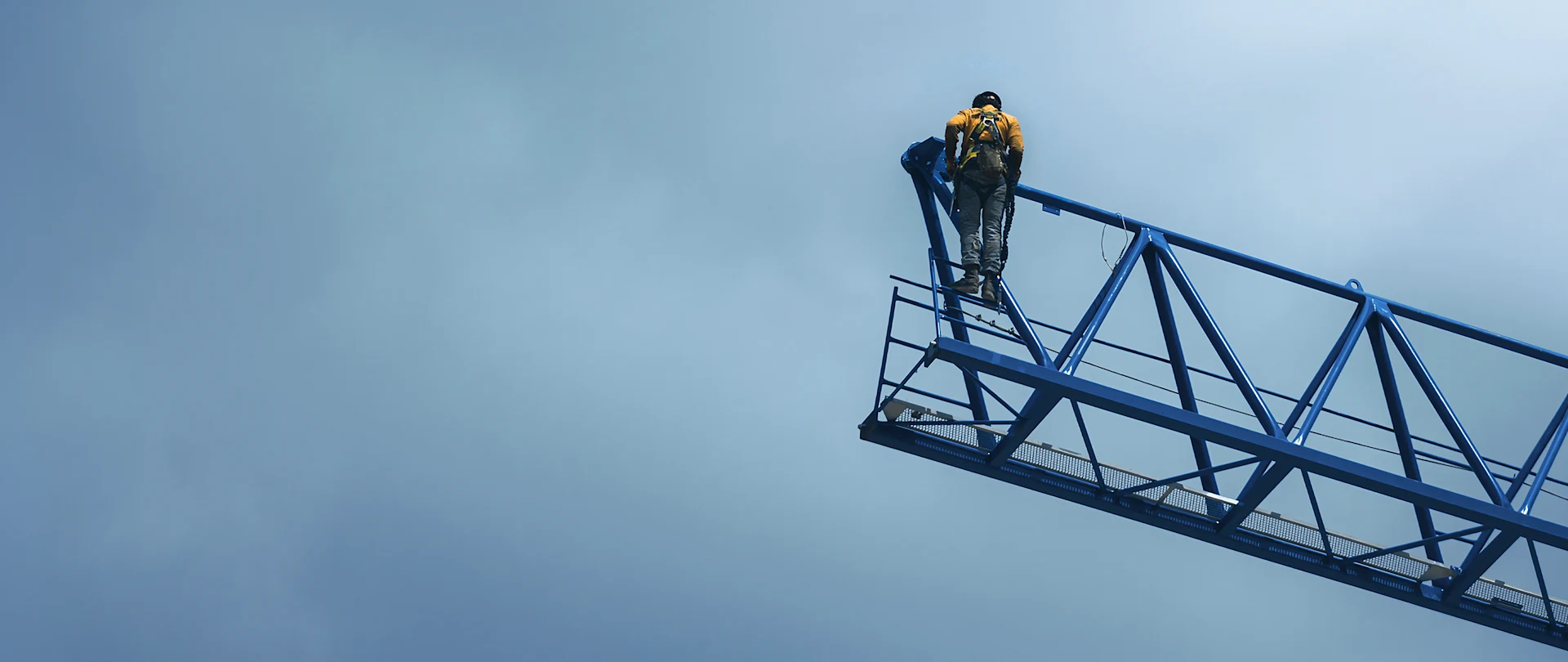 A construction worker in safety gear stands on a blue crane jib against a cloudy sky, representing Trimble construction technology.