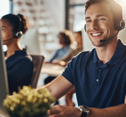 Smiling man wearing a headset works at a computer in a bright office. Colleagues in headsets are in the background