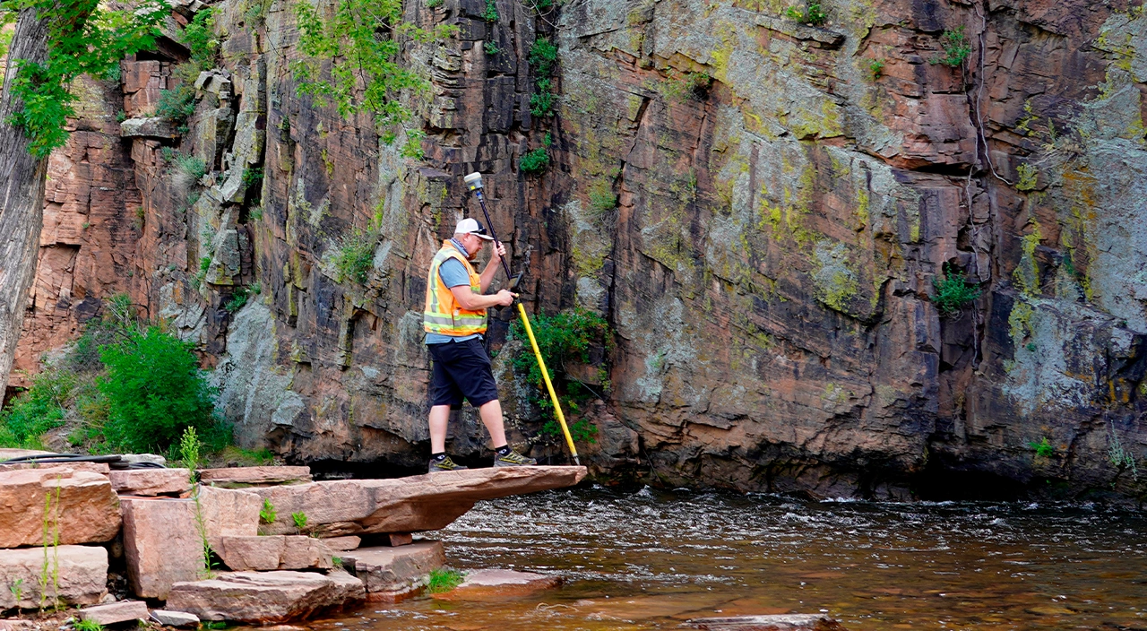 Eine Person in einer Sicherheitsweste steht auf einem Felsvorsprung über einem Fluss und benutzt Trimble-Vermessungsgeräte.