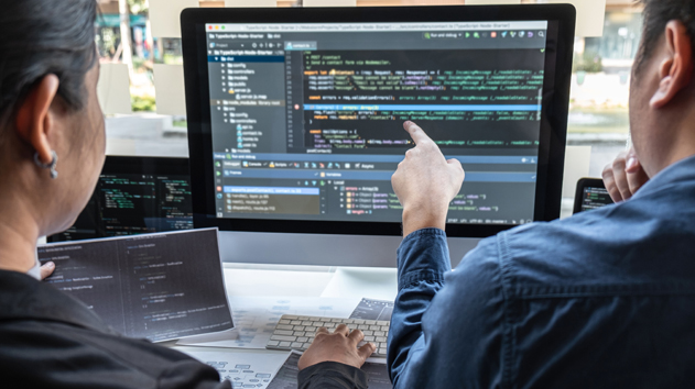 Two people collaborate at a desk, focusing on a large monitor displaying complex code with one pointing at the screen.