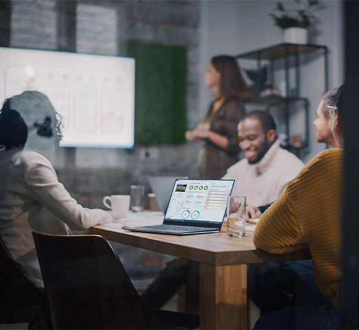 Office meeting with a laptop displaying graphs, people seated around a table, and a presentation screen.